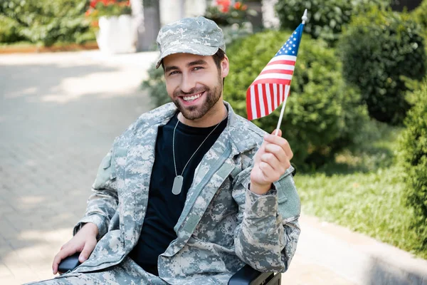 Veterano discapacitado en uniforme militar sosteniendo pequeña bandera de EE.UU. y sonriendo en la calle de la ciudad - foto de stock