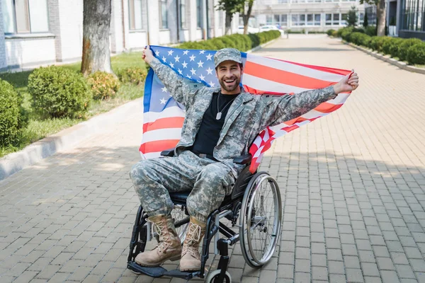 Veterano desativado feliz olhando para a câmera enquanto sentado em cadeira de rodas com bandeira dos EUA — Fotografia de Stock