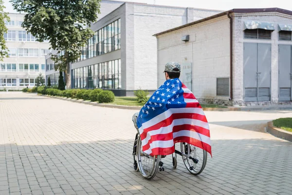 Back view of young veteran, covered with usa flag, sitting in wheelchair in city — Stock Photo