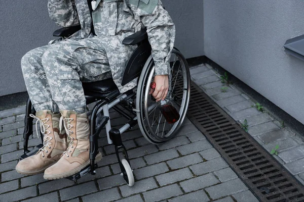 Cropped view of disabled soldier sitting in wheelchair and holding bottle of whiskey outdoors — Stock Photo
