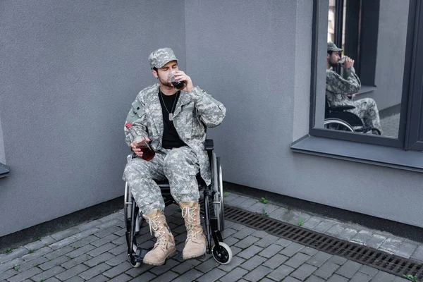 Young disabled soldier in wheelchair drinking alcohol in corner of grey wall outdoors — Stock Photo