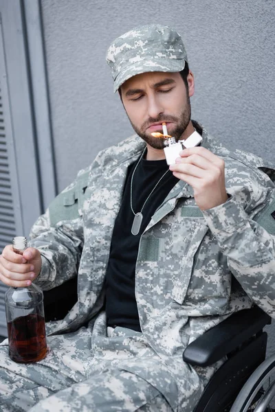 Jovem soldado deficiente acendendo cigarro enquanto segurando garrafa de álcool ao ar livre — Fotografia de Stock