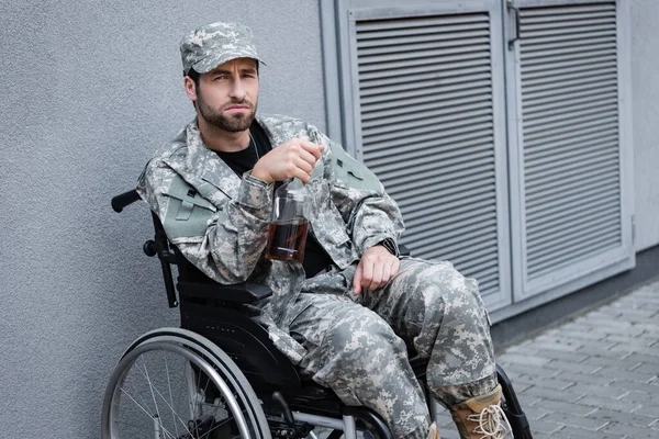 Militar deprimido y discapacitado sentado en silla de ruedas con botella de alcohol - foto de stock