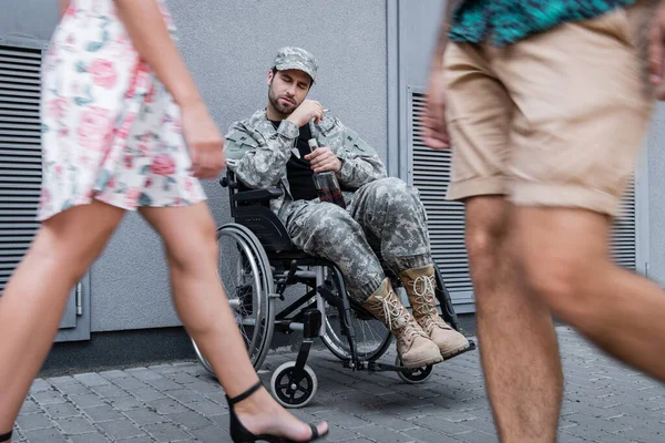 Drunk disabled soldier sitting in wheelchair with bottle and cigarette near blurred strangers passing by — Stock Photo