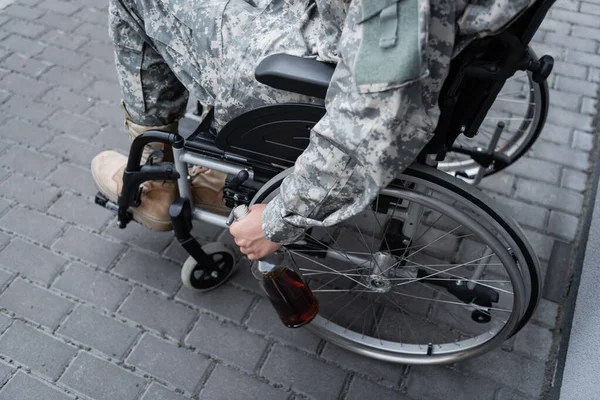 Partial view of handicapped military man holding bottle of alcohol while sitting in wheelchair outdoors — Stock Photo