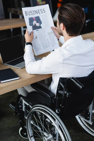 Jeune homme d'affaires handicapé dans un journal de lecture en fauteuil roulant près d'un ordinateur portable avec écran blanc — Photo de stock