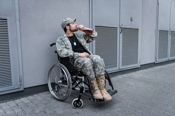 Jovem veterano deficiente sentado em cadeira de rodas na rua da cidade e bebendo uísque — Fotografia de Stock