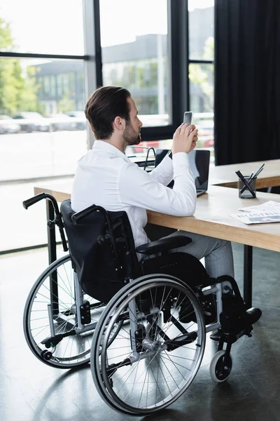 Uomo d'affari disabile in sedia a rotelle messaggistica su smartphone vicino al computer portatile in ufficio — Foto stock