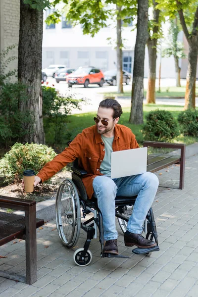 Smiling disabled man holding coffee to go while sitting in wheelchair with laptop — Stock Photo