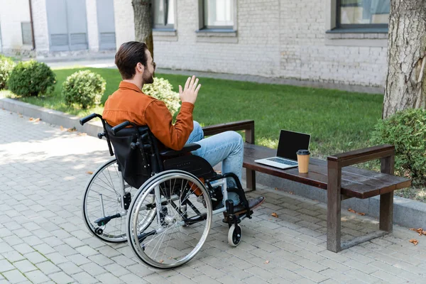 Uomo handicappato in sedia a rotelle mano agitando durante la video chat sul computer portatile all'aperto — Foto stock