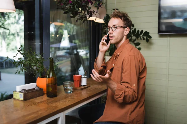 Joven en gafas haciendo gestos mientras habla en el teléfono inteligente en la cafetería — Stock Photo
