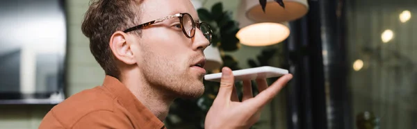 Young man in glasses recording voice message on smartphone in cafe, banner — Stock Photo