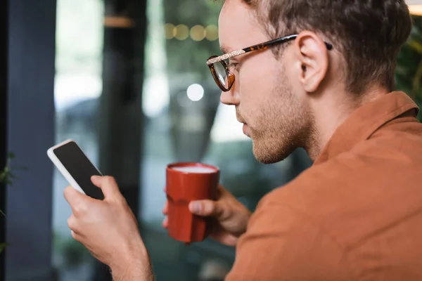 Young man in glasses holding cup of coffee and texting on smartphone in cafe — Stock Photo