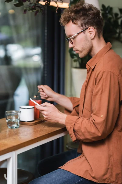 Blurred young man in glasses holding spoon and texting on smartphone in cafe — Stock Photo