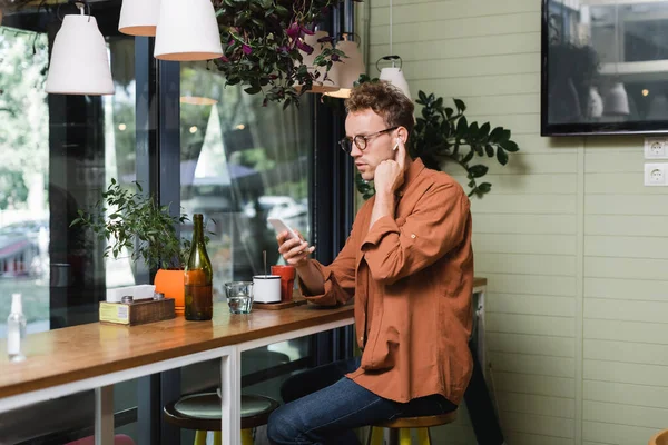 Jovem em óculos ajustando fones de ouvido sem fio e usando smartphone no café — Fotografia de Stock