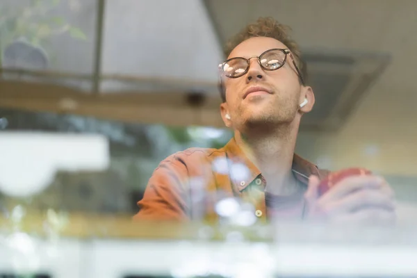 Vue à angle bas de l'homme dans des lunettes et écouteurs sans fil tenant tasse derrière la fenêtre floue du café — Photo de stock