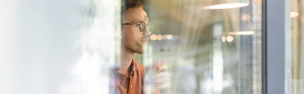 Young man in glasses and wireless earphones holding cup of coffee behind blurred window of cafe, banner — Stock Photo