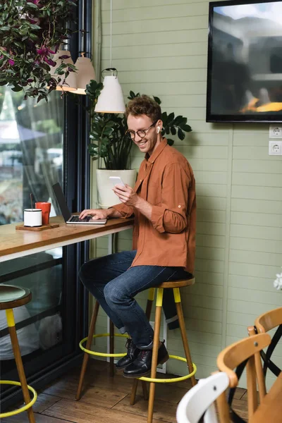 Happy young freelancer in glasses using smartphone near laptop in cafe — Stock Photo