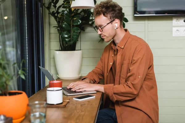 Молодий фрілансер в окулярах, що друкують на ноутбуці біля смартфона в кафе — стокове фото
