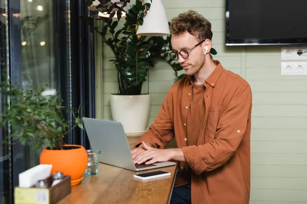 Jeune pigiste dans les lunettes de dactylographie sur ordinateur portable près du téléphone cellulaire dans le café — Photo de stock