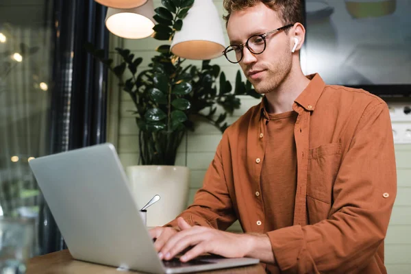 Freelancer in glasses and wireless earphones typing on blurred laptop in cafe — Stock Photo