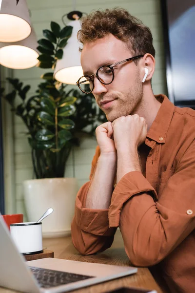 Jovem freelancer em óculos e fones de ouvido sem fio olhando para laptop borrado no café — Fotografia de Stock