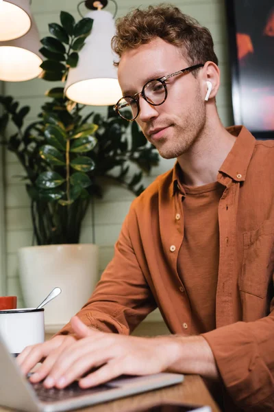 Young freelancer in glasses and wireless earphones typing on blurred laptop in cafe — Stock Photo