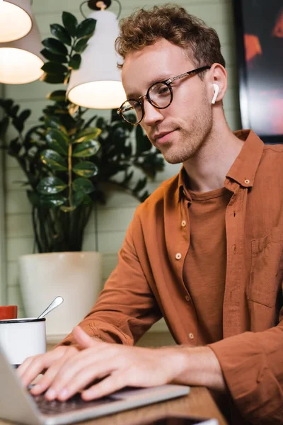 Giovane freelance in occhiali e auricolari wireless digitando sul computer portatile offuscata in caffè — Foto stock