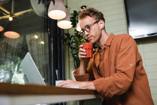 Young freelancer in glasses and wireless earphones holding cup and using laptop in cafe — Stock Photo