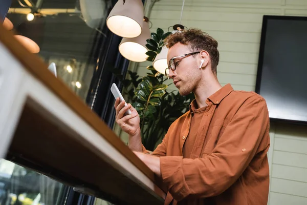 Low angle view of young man in glasses and wireless earphones using smartphone in cafe — Stock Photo