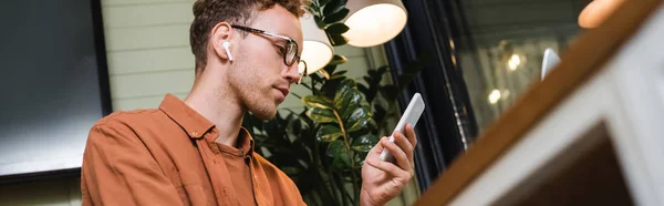 Low angle view of young man in glasses and wireless earphones using smartphone in cafe, banner — Stock Photo