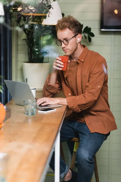 Joven freelancer en gafas sosteniendo la taza y escribiendo en el portátil cerca de teléfono inteligente en la cafetería - foto de stock