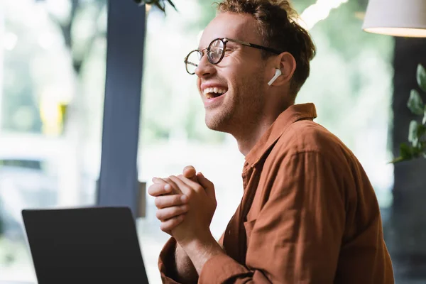 Freelancer feliz em óculos e fones de ouvido sentado com as mãos apertadas perto de laptop com tela em branco no café — Fotografia de Stock