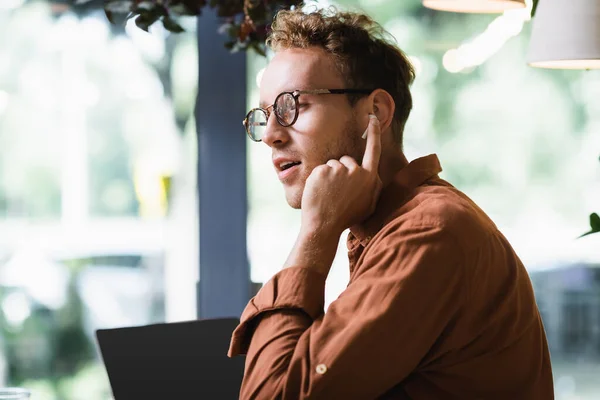 Young freelancer in glasses adjusting earphones near laptop with blank screen in cafe — Stock Photo