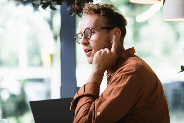 Joven freelancer en gafas ajustando auriculares cerca de laptop con pantalla en blanco en cafetería - foto de stock