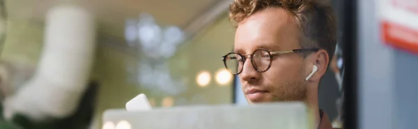 Jeune homme dans des lunettes et écouteurs près d'un ordinateur portable derrière la fenêtre floue du café, bannière — Photo de stock