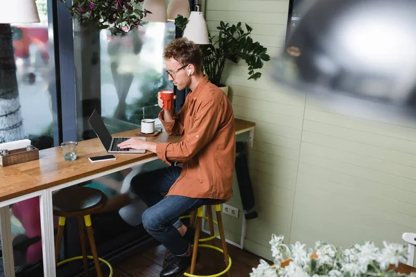 Freelancer en audífonos sujetando taza y usando laptop cerca del celular en la mesa en la cafetería - foto de stock