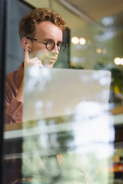 Young man in glasses adjusting earphones near laptop behind blurred window of cafe — Stock Photo