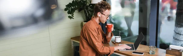 Freelancer em fones de ouvido segurando copo e usando laptop perto de smartphone na mesa no café, banner — Fotografia de Stock