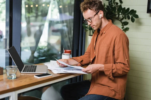 Freelancer in earphones looking at graphs near gadgets and drinks on table in cafe — Stock Photo