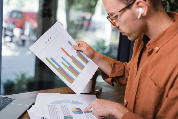 Young man in glasses and earphones looking at graphs near laptop in cafe — Stock Photo