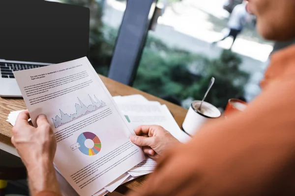 Vista recortada de hombre joven sosteniendo gráficos y gráficos cerca de la computadora portátil en la cafetería — Stock Photo