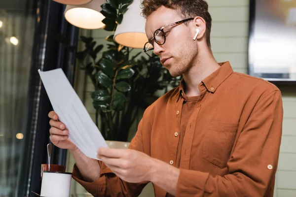 Giovane in occhiali e auricolari guardando carta con grafici in caffè — Foto stock