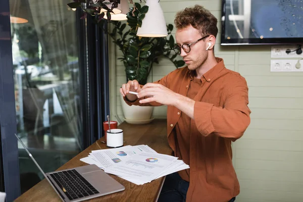Freiberufler mit Kopfhörer und Brille, der im Café Fotos von Grafiken in der Nähe von Laptops macht — Stockfoto