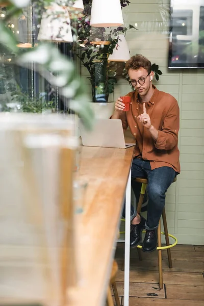 Homme en lunettes et écouteurs sans fil pointant du doigt tout en ayant un appel vidéo sur ordinateur portable — Photo de stock