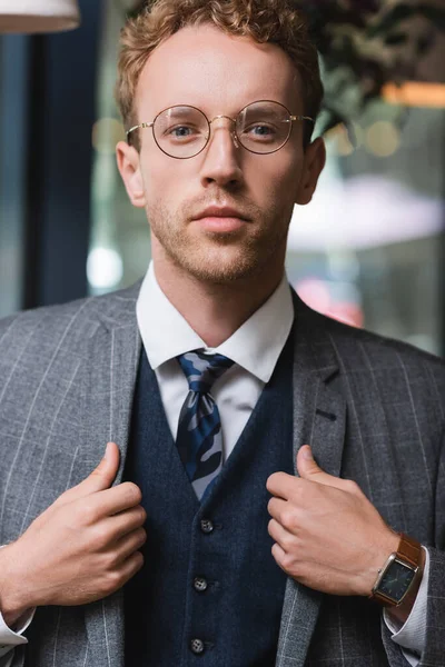 Joven hombre de negocios guapo en ropa formal y gafas ajustando chaqueta en la cafetería - foto de stock