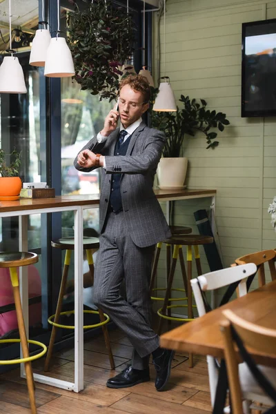 Longueur complète de jeune homme d'affaires en tenue formelle regardant montre-bracelet tout en parlant sur smartphone dans le café — Photo de stock