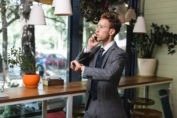 Giovane uomo d'affari in abito formale con orologio da polso a portata di mano parlando su smartphone in caffè — Foto stock
