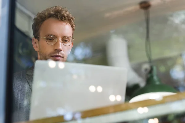 Кудрявый бизнесмен в очках с ноутбуком за размытым окном кафе — стоковое фото