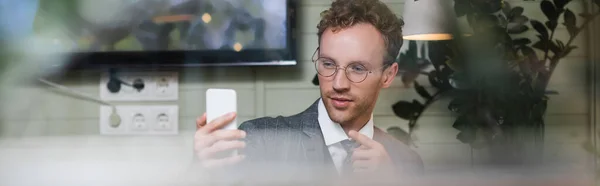 Hombre de negocios rizado en traje apuntando con el dedo mientras toma selfie detrás de ventana borrosa de la cafetería, pancarta - foto de stock